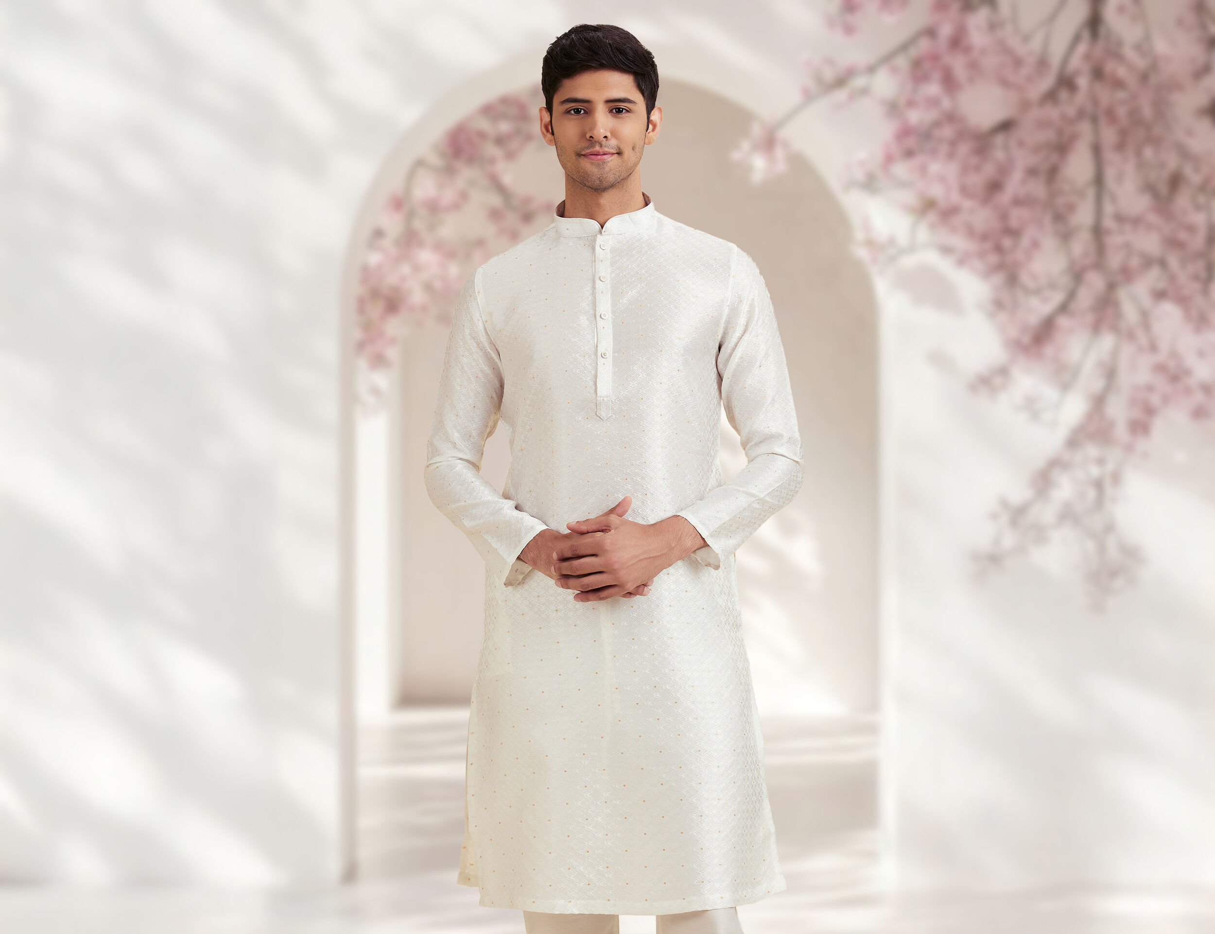 Man in cream embroidered silk kurta posing against an elegant arch backdrop with blooming cherry blossoms—traditional Onam men’s attire 2025.